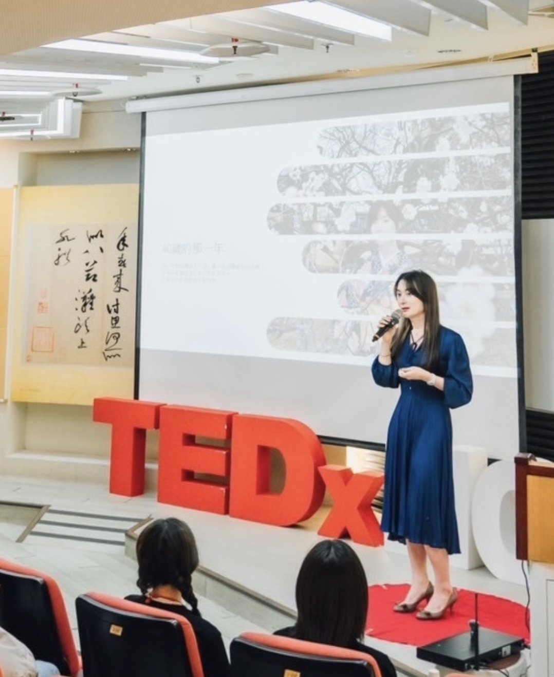 陳樂涵身穿藍色洋裝，手持麥克風在 TEDx 舞台上分享的演講現場